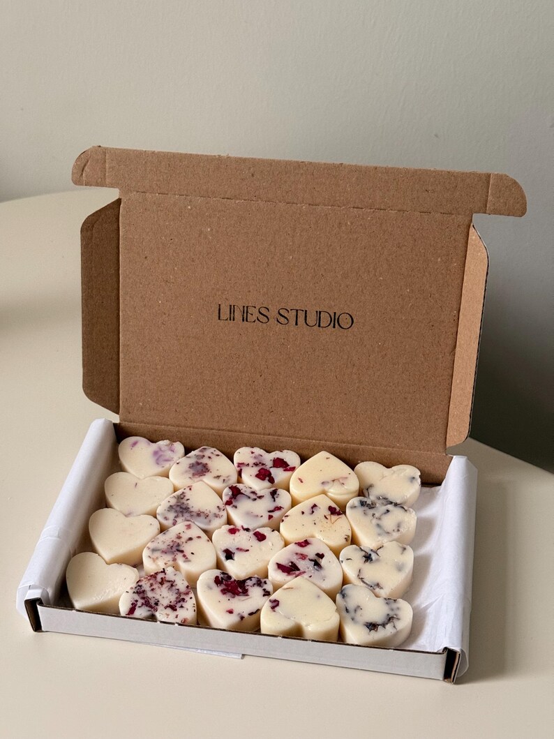 May include: A cardboard box with the text "LINES STUDIO" on the lid, open to reveal heart-shaped wax melts. Some melts are plain, while others are decorated with dried flowers. The box is lined with white paper.