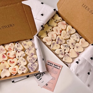 May include: Two open cardboard boxes filled with heart-shaped wax melts. The melts are white with floral accents in pink and purple. The boxes sit on white tissue paper with black heart patterns. A pink card with text is visible.