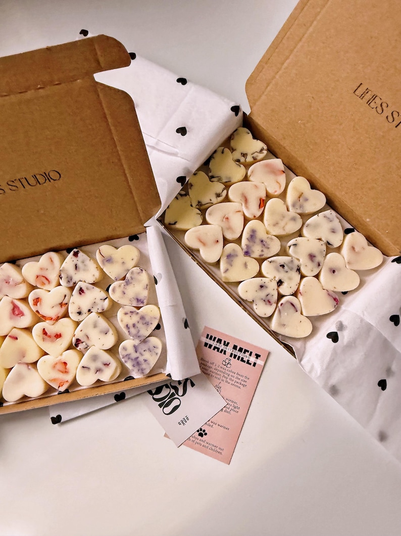 May include: Two open cardboard boxes filled with heart-shaped wax melts. The melts are white with floral accents in pink and purple. The boxes sit on white tissue paper with black heart patterns. A pink card with text is visible.