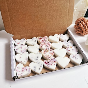 May include: A box of heart-shaped wax melts. Some melts are plain white, while others are decorated with dried flowers and herbs. The box is open, revealing the contents, and is made of brown cardboard. The melts are arranged neatly inside.