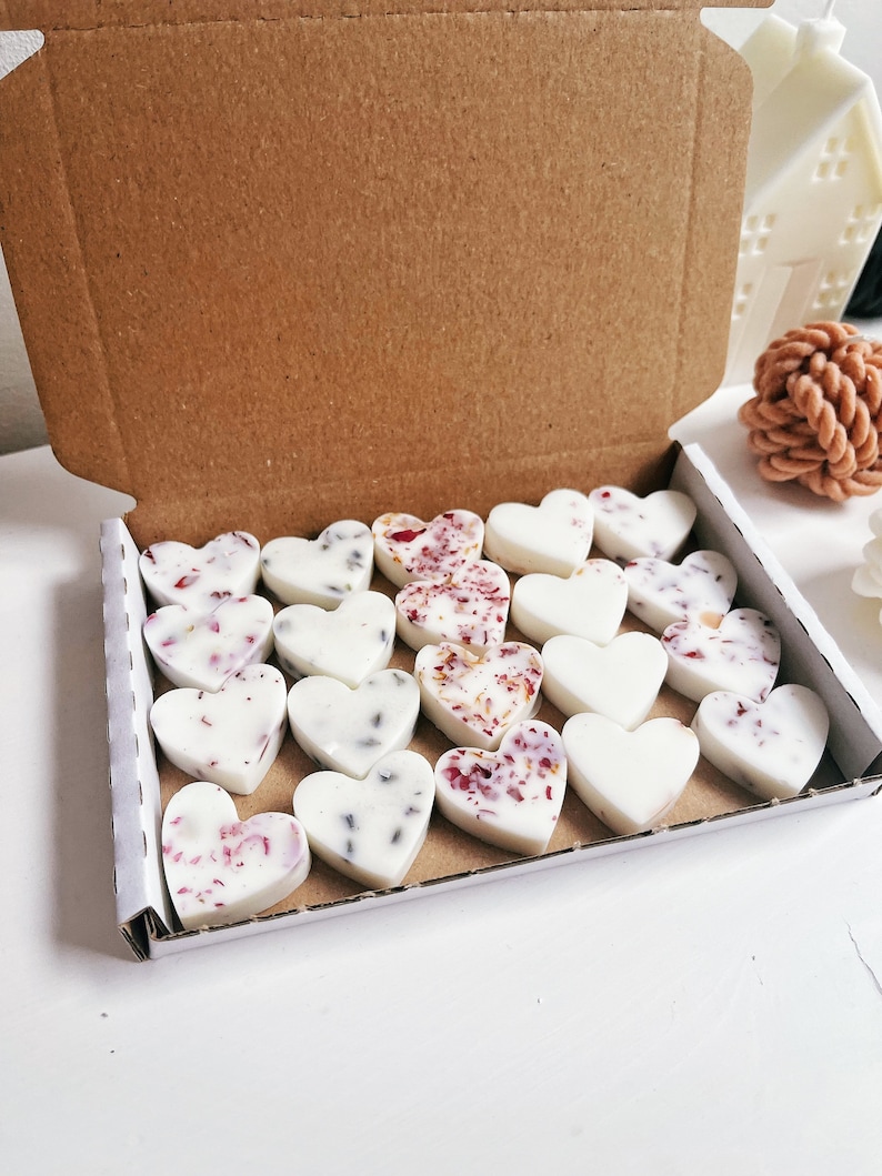 May include: A box of heart-shaped wax melts. Some melts are plain white, while others are decorated with dried flowers and herbs. The box is open, revealing the contents, and is made of brown cardboard. The melts are arranged neatly inside.