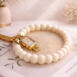 May include: A gold wristwatch with a rectangular face rests on a white beaded bracelet. The watch and bracelet are on a book with the words "TRAVEL & STYLE" visible. The background includes white flowers in a vase.