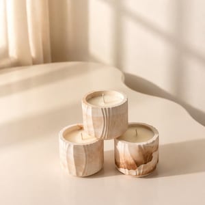 May include: Three small, round candles in beige and brown textured holders. The candles are stacked on a light-colored surface, with a soft, neutral background. The holders have a carved, organic design.