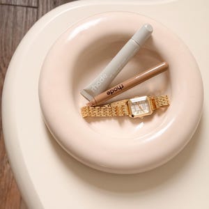 Può includere: Un orologio da polso dorato con quadrante rettangolare e cinturino a maglie è appoggiato in una ciotola circolare rosa chiaro. Due tubetti di trucco del marchio Rhode, uno grigio e uno marrone, sono anch'essi nella ciotola. La ciotola è su una superficie color crema.