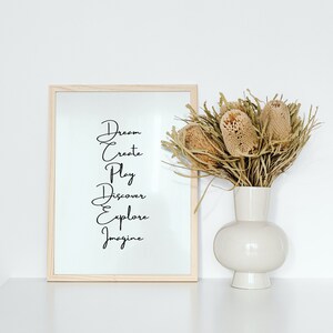 May include: A framed print with the words "Dream, Create, Play, Discover, Explore, Imagine" in black script on a white background. The print is sitting on a white surface next to a vase with dried flowers.