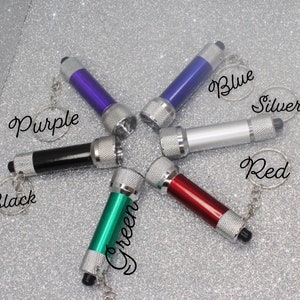 Może przedstawiać: Sześć latarek breloczków w różnych kolorach: fioletowy, czarny, zielony, czerwony, niebieski i srebrny. Każda latarka ma srebrny metalowy pierścień i srebrny metalowy przycisk.