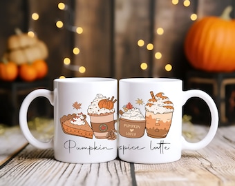 May include: Two white ceramic mugs featuring colorful illustrations of pumpkin pie, whipped cream, and coffee drinks. The text "Pumpkin spice latte" is printed in black script across the front of the mugs, accompanied by small autumn leaf and pinecone graphics.