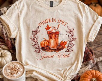 May include: A cream-colored t-shirt featuring a graphic of a glass mug filled with a pumpkin spice beverage, topped with whipped cream and a red bow. The text reads PUMPKIN SPICE Social Club, surrounded by orange pumpkins and autumn leaves.