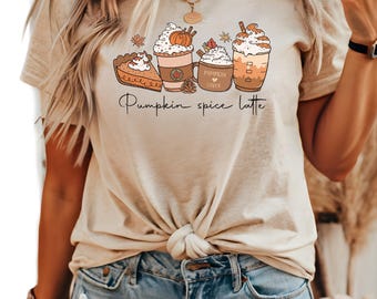 May include: A beige short-sleeve t-shirt featuring a graphic print of a slice of pumpkin pie, two coffee cups with whipped cream, and a tall iced coffee. The text "Pumpkin spice latte" is printed in black cursive script below the illustrations.