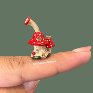 Puede incluir: Una figura miniatura de cerámica en forma de casa de hongo con un sombrero rojo brillante con puntos blancos, una base beige con ventanas y puerta pintadas, y una extensión curva similar a una chimenea.