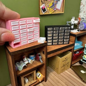 May include: Miniature pink storage cabinet with white drawers, next to a black cabinet with clear drawers. The cabinets sit on a wooden desk with various miniature tools and supplies. A corkboard with small pictures is in the background.