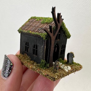 Może przedstawiać: Miniaturowy czarny drewniany domek z dachem pokrytym mchem i małym drzewem z przodu. Domek ma dwa okna i drzwi. Przed domkiem znajduje się mały cmentarz z nagrobkiem.