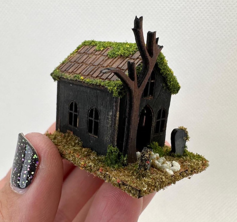 Może przedstawiać: Miniaturowy czarny drewniany domek z dachem pokrytym mchem i małym drzewem z przodu. Domek ma dwa okna i drzwi. Przed domkiem znajduje się mały cmentarz z nagrobkiem.