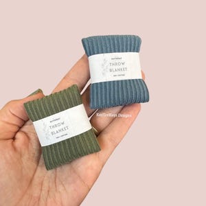 Op de afbeelding: Twee kleine, opgerolde sprei, één olijfgroen en één blauwgrijs, worden in een hand gehouden. Elke deken is verpakt met een witte papieren band met de tekst "THROW BLANKET" en "100% COTTON". De dekens zijn geribbeld.