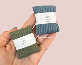 May include: Two small, rolled throw blankets, one olive green and one blue-grey, are held in a hand. Each blanket is wrapped with a white paper band that reads "THROW BLANKET" and "100% COTTON". The blankets are ribbed.