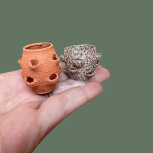 May include: Two small, handmade strawberry planters. One is terracotta-colored with multiple openings, and the other is speckled gray and brown. The planters are held in a hand against a green background. The text "KayTeeKays Designs" is visible.