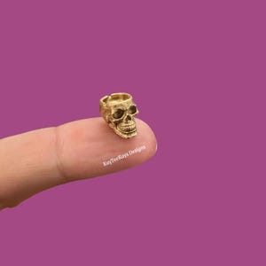 May include: A small, gold-colored skull figurine with detailed facial features. The skull is resting on a finger against a purple background. The text "KayTeeKays Designs" is visible.