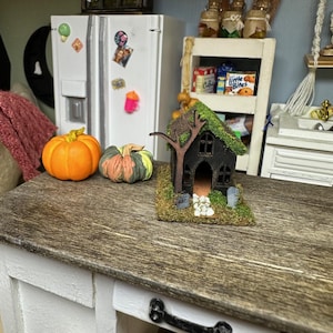 Może przedstawiać: Miniaturowy dom w stylu Halloween z czarnym zewnętrzem, zielonym mchem na dachu i białym płotkiem. Dom otoczony jest białymi kamieniami i stoi na drewnianym stole z dwoma pomarańczowymi dyniami.