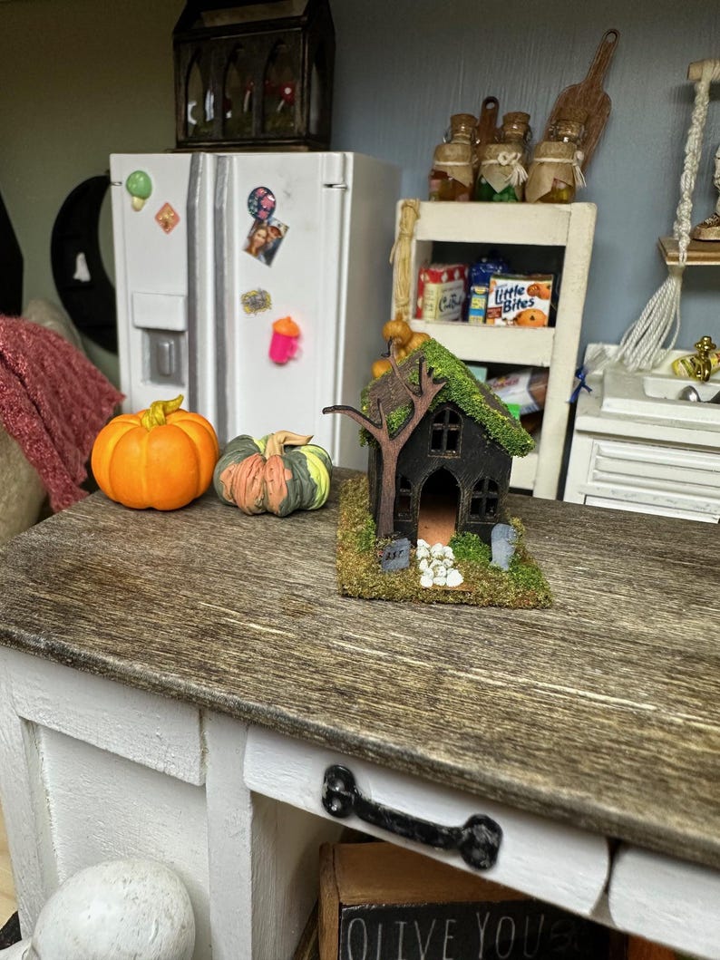 Może przedstawiać: Miniaturowy dom w stylu Halloween z czarnym zewnętrzem, zielonym mchem na dachu i białym płotkiem. Dom otoczony jest białymi kamieniami i stoi na drewnianym stole z dwoma pomarańczowymi dyniami.