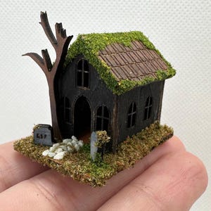 Może przedstawiać: Miniaturowy drewniany dom z duchami z dachem pokrytym mchem, krzywym drzewem i małym nagrobkiem z napisem "RIP".