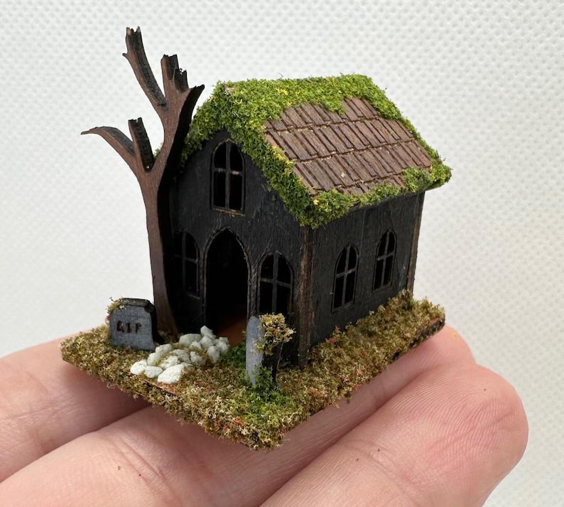 Może przedstawiać: Miniaturowy drewniany dom z duchami z dachem pokrytym mchem, krzywym drzewem i małym nagrobkiem z napisem "RIP".