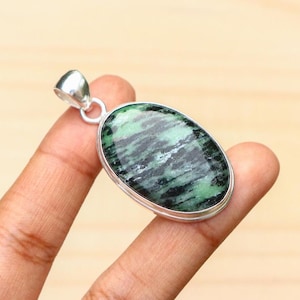May include: Oval pendant featuring a green and black stone, set in a silver-coloured metal frame. The stone has a marbled effect with shades of green and black. The pendant is suspended from a small loop at the top.