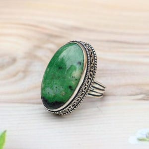 Puede incluir: Un anillo de plata con una gran piedra verde de forma ovalada con inclusiones oscuras. La banda de metal está decorada con un patrón detallado y repetitivo de volutas en los bordes, que enmarca la superficie pulida y lisa de la gema.