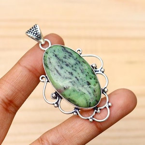 May include: A silver pendant with an oval, green and black stone. The stone is set in a decorative silver frame with a loop at the top for a chain. The pendant is held by a hand.