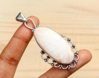 Può includere: Ciondolo in argento con una pietra ovale bianco sporco con motivo strutturato. La mano tiene il ciondolo, evidenziando l'incastonatura in argento e l'anello decorativo. Il motivo della pietra ricorda il corallo.