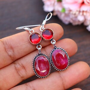 Puede incluir: Un par de pendientes de plata con detalles de piedras preciosas rojas. Los pendientes tienen dos piedras preciosas rojas, una redonda y una ovalada, engastadas en plata.