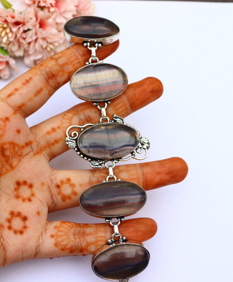May include: A silver bracelet featuring four oval-shaped fluorite gemstones. The fluorite is a deep purple with streaks of white and grey. The bracelet is made of sterling silver and has a delicate design.