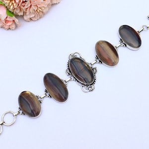 May include: A silver bracelet featuring five oval-shaped iridescent stones. The stones display a blend of brown, grey, and black hues. The bracelet has a toggle clasp.