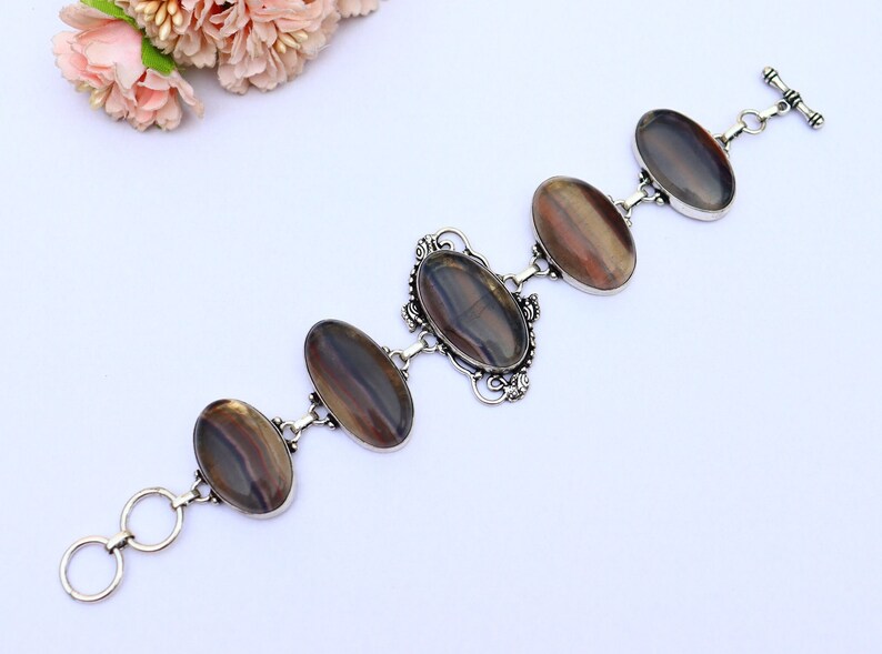 May include: A silver bracelet featuring five oval-shaped iridescent stones. The stones display a blend of brown, grey, and black hues. The bracelet has a toggle clasp.