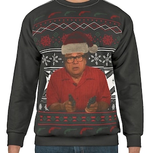 May include: A black sweatshirt with a Christmas sweater design featuring a man wearing a Santa hat and holding two guns. The sweater has a red, green, and white pattern with snowflakes and guns.