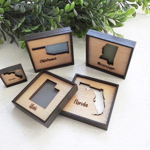 May include: Five wooden state outline signs with black frames. The states are Florida, Utah, Oklahoma, Mississippi, and Nevada. Each sign has the state name written below the outline.