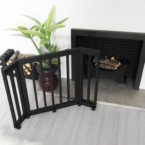 May include: Black metal fireplace screen with a decorative design. The screen is folded and sitting on a light wood floor in front of a fireplace.