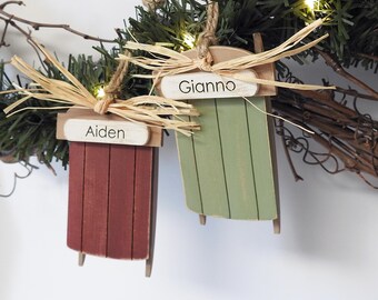 May include: Two wooden sled ornaments, one red and one green, are suspended from a branch. Each sled has a name tag with a name printed on it. The red sled says "Aiden" and the green sled says "Gianno". The ornaments are decorated with raffia.