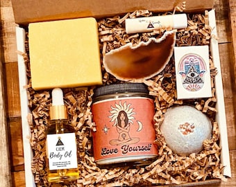 May include: A brown box filled with self-care items. The box has a logo that says "Under the Seventh Ray". The items include a bar of soap, a bottle of body oil, a candle, a bath bomb, a lip balm, a small agate slice, and a sticker with a hamsa hand design.
