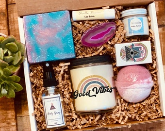 May include: A gift box filled with self-care items, including a pink and blue soap bar, a candle with the text "Good Vibes", a bath bomb, a lip balm, a body spray, and a small box with a hamsa hand design.