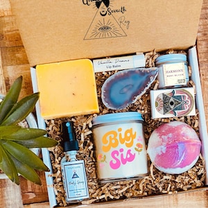 May include: A gift box filled with self-care items, including a bar of soap, a lip balm, a body butter, a bath bomb, a candle, a body spray, and a crystal. The candle has the text "Big Sis" on it.