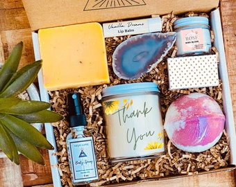 May include: A gift box filled with self-care items, including a bar of soap, a candle, a bath bomb, a lip balm, a body spray, and a small jar of rose body butter. The box is decorated with a brown paper and a tag that says "Thank You".
