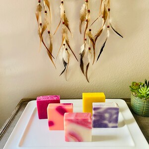 May include: Five bars of handmade soap on a white rectangular plate. The soaps are in various colors and patterns, including pink, purple, yellow, and white.