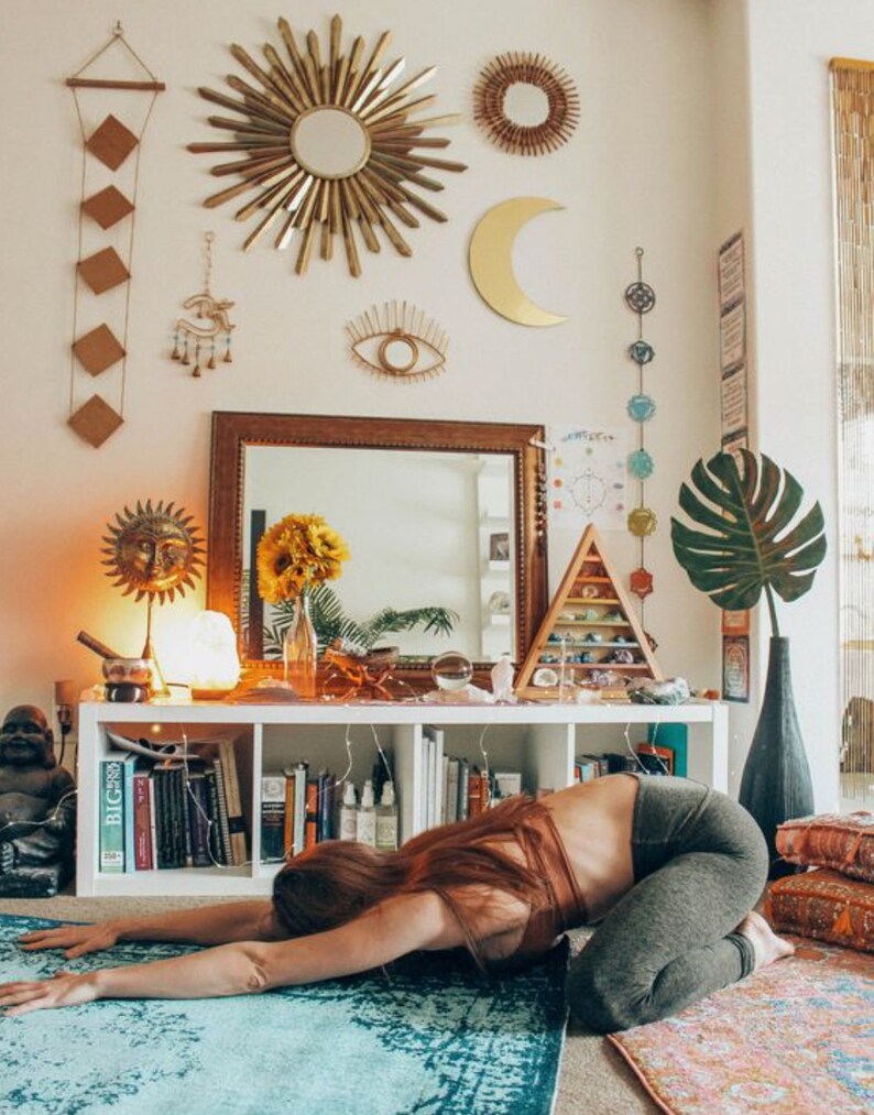 May include: A woman in a brown tank top and gray leggings is doing a yoga pose on a blue and white rug. The room is decorated in a bohemian style with a mirror, a sun and moon wall decor, and a shelf with crystals and books.