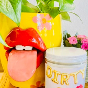Peut inclure: Un pot de fleurs en c&eacute;ramique jaune avec un design de l&egrave;vres rouges et une langue tir&eacute;e, contenant des plantes vertes. Un pot de bougie avec le texte "DIRTY 30" en lettres dor&eacute;es et un motif de l&egrave;vres rouges est &eacute;galement pr&eacute;sent.