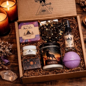 May include: An open cardboard gift box filled with various self-care items. Includes a bar of soap, a jar of cream, a candle with an Aries design, a spray bottle, a bath bomb, and a small bag. The box is surrounded by lit candles.