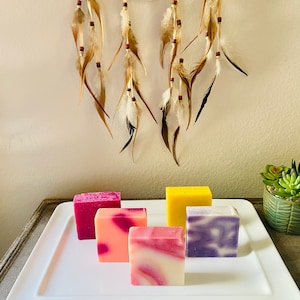 May include: Five bars of handmade soap on a white rectangular plate. The soaps are in various colors and patterns, including pink, purple, yellow, and white.