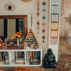 May include: A white bookshelf with books and crystals, a black Buddha statue, and a rug with an orange, brown, and white pattern. The wall behind the bookshelf has a crescent moon, a wall hanging with an eye, and a string of chakra symbols.