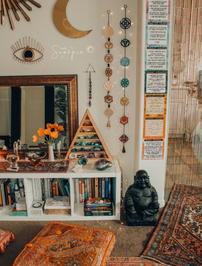 May include: A white bookshelf with books and crystals, a black Buddha statue, and a rug with an orange, brown, and white pattern. The wall behind the bookshelf has a crescent moon, a wall hanging with an eye, and a string of chakra symbols.
