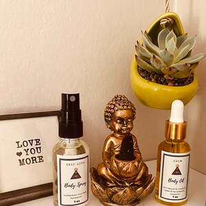May include: Two small bottles with gold lids and labels. The bottles are labeled "Self-Love Body Spray" and "Calm Body Oil". The bottles are sitting on a table with a small golden Buddha statue and a succulent plant in a yellow pot.
