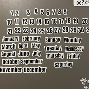 May include: A set of black and white magnetic calendar pieces featuring the numbers 1 through 31, the twelve months of the year, and the seven days of the week. Each piece has a bold white outline and clear sans-serif typography for display.